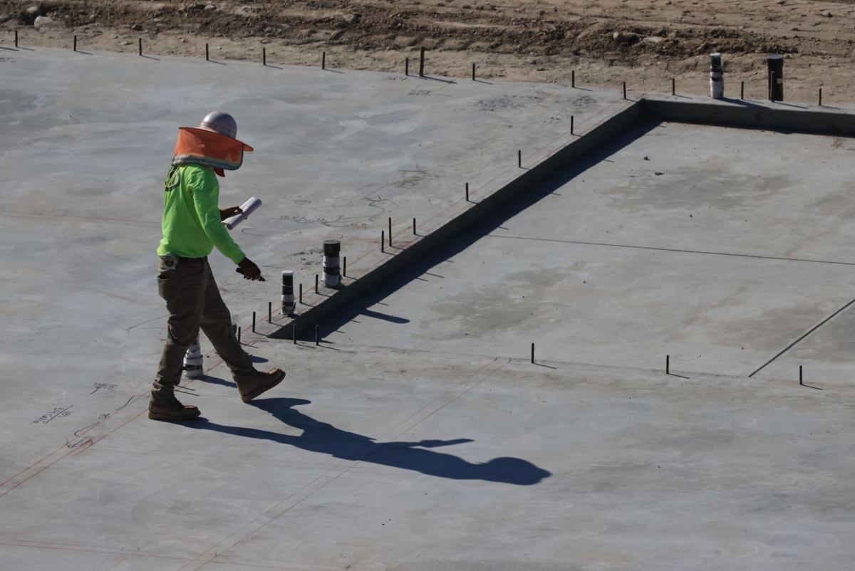 Engineer in a hard hat walking a freshly poured concrete slab foundation with plumbing stub-ups, checking the work